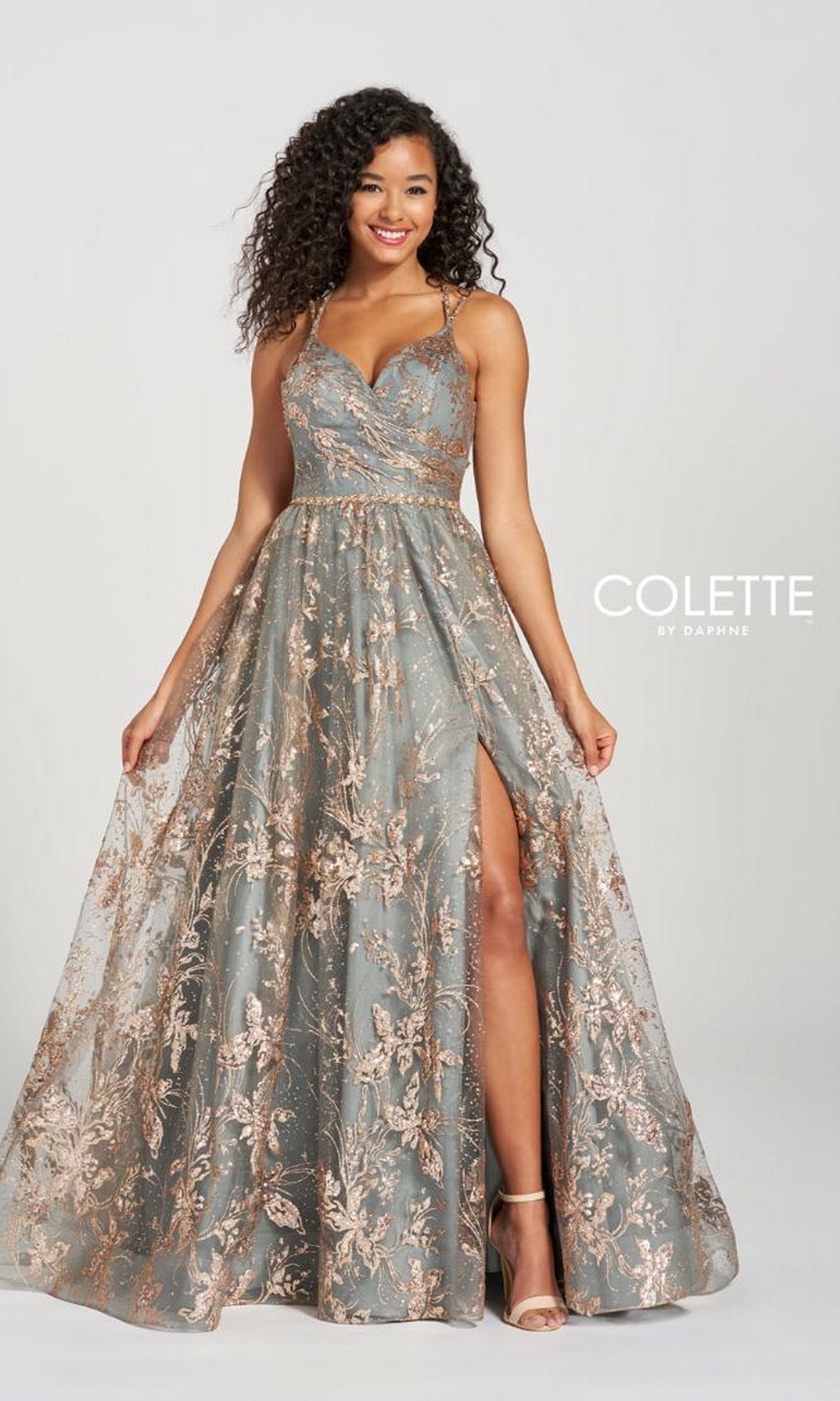 A woman poses against a plain background in the Gold and Pewter Prom Ball Gown Colette CL12218, featuring a sleeveless design, long A-line silhouette, floral lace overlay, fitted bodice, and a thigh-high slit.
